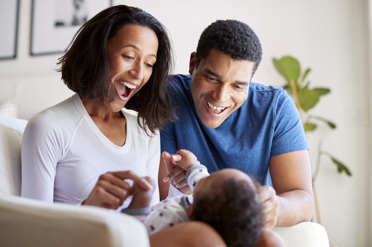 Happy mixed race young adult parents playing with their three month old baby son, lying on his motherâ"u20acs knee, selective focus