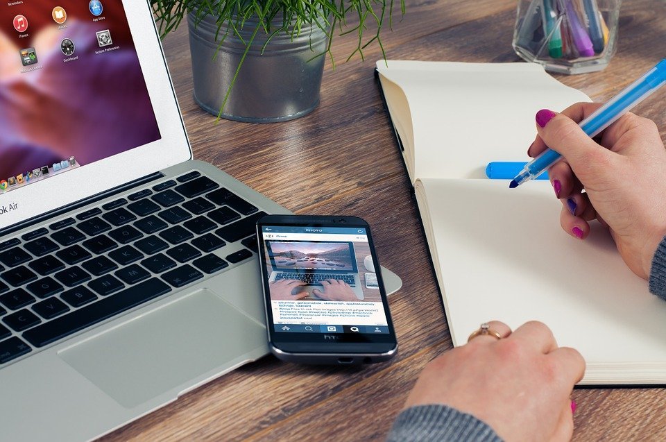 woman writing on a notebook beside a laptop and a phone on a wooden table