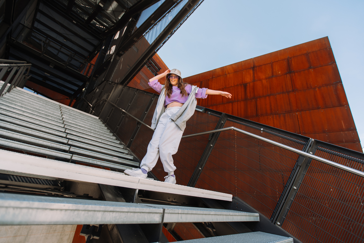Joyful caucasian girl in hip hop outfit wearing dancing into music under street. Urban background. Wide shot