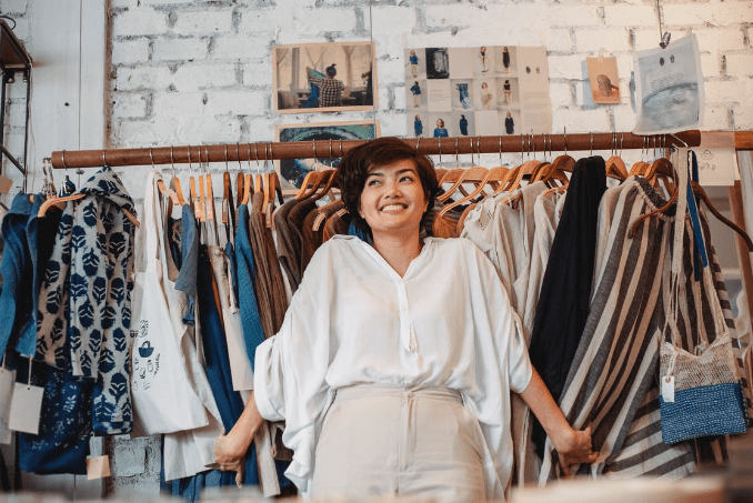 cheerful-young-ethnic-lady-smiling-while-holding-stack-of-clothes-hanging-on-rack