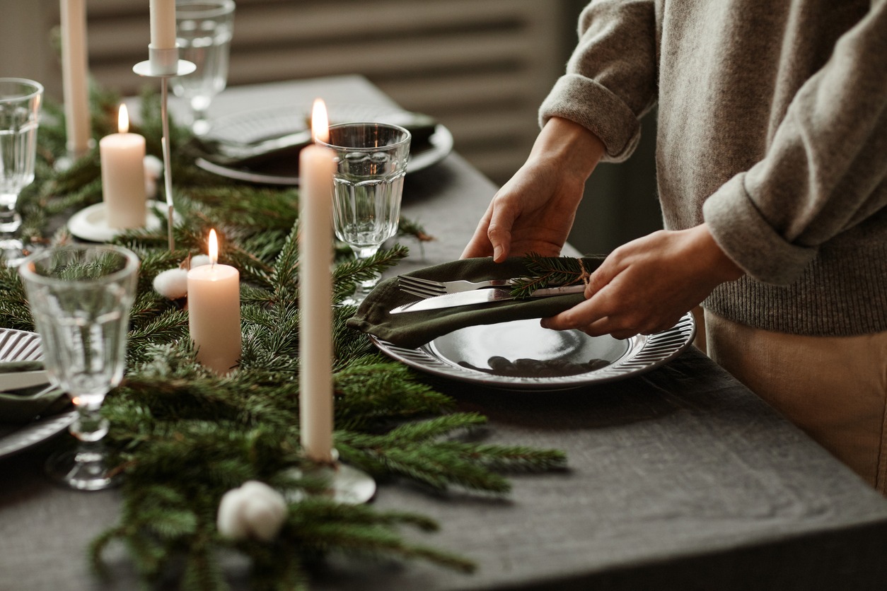 Side view close up of unrecognizable woman setting up dining table decorated for Christmas with fir branches and candles in grey tones