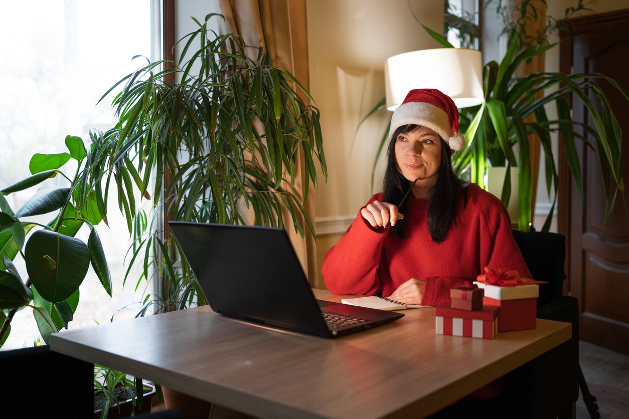 Thoughtful young woman holding pencil in hand on chin and looking away while sitting at table eve Christmas